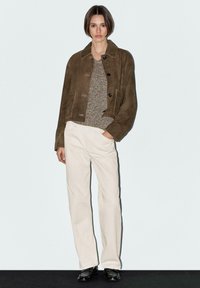 Brown suede jacket with a collared neckline, two pockets, and button closure, worn over a textured sweater and cream-colored trousers.