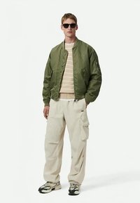 Άνδρας που φοράει σκούρα γυαλιά ηλίου, πράσινο bomber μπουφάν, μπεζ ριγέ πουλόβερ, κρεμ cargo παντελόνι και αθλητικά παπούτσια, στέκεται σε λευκό φόντο.