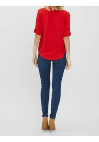 Rød kortærmet bluse med optrillede ærmer, parret med mørkeblå skinny jeans og lysebrune højhælede sko. Blød tekstur, afslappet design.