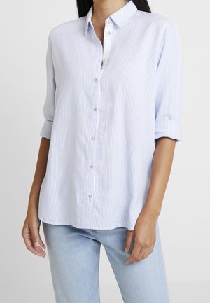 Frau trägt ein hellblau-weiß vertikal gestreiftes Hemd mit hochgekrempelten Ärmeln und eine hellblaue Jeans vor einem einfarbigen Hintergrund.