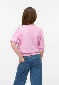 Ragazza con lunghi capelli castani che indossa un maglione rosa e jeans blu, in piedi con una mano nella tasca posteriore contro uno sfondo bianco.