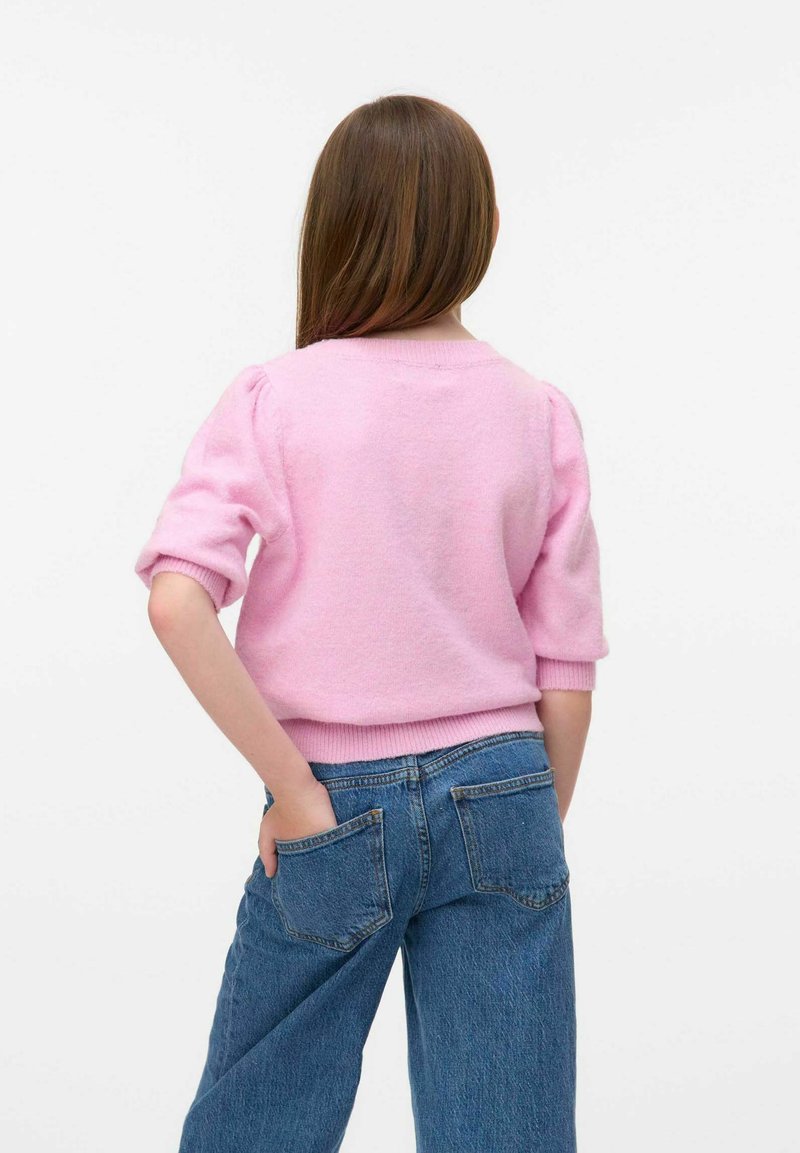 Ragazza con lunghi capelli castani che indossa un maglione rosa e jeans blu, in piedi con una mano nella tasca posteriore contro uno sfondo bianco.