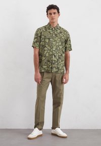 Korte mouwen groene bloemenoverhemd met een zwart patroon, knoopsluiting aan de voorkant, gecombineerd met beige broeken en witte sneakers, staand op een grijze vloer.