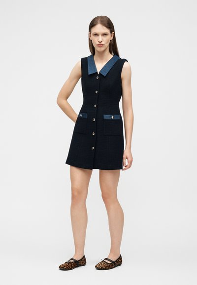 Γυναίκα όρθια, φορώντας μαύρο φόρεμα χωρίς μανίκια με κουμπιά μπροστά, μπλε γιακά και διακοσμητικές τσέπες, συνδυασμένο με παπούτσια leopard print.