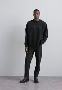 Must mustaga, millel on peen teksti disain, koos tumehalli pükste ja mustade nahast saapatega, seistes neutraalse tausta ees.