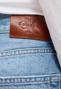 Καφέ δερμάτινο μπαλώμα Calvin Klein με το λογότυπο στη ζώνη μιας ανοικτόχρωμης μπλε τζιν παντελόνι κάτω από ένα λευκό πουκάμισο.