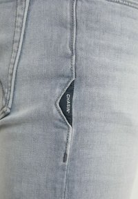 Närbild av ljusgrå denimjeansficka med sydd söm och liten svart tygetikett med texten "CHASIN'".