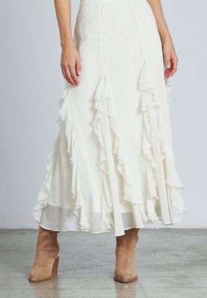 Weißer Midirock mit mehrlagigen Rüschen, kombiniert mit beige Wildleder-Stiefeletten auf grauem Boden.