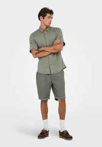 Chemise à manches courtes olive avec un design à boutons; associée à des shorts gris retroussés. Des chaussures brunes et des chaussettes blanches complètent le look.