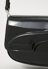 Crna lakirana kožna torba preko tijela s zakrivljenim flapom, sjajna površina, srebrni dodaci i kontrastni detalj šivanja.