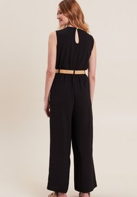 Zwarte mouwloze jumpsuit met een keyhole-opening op de rug, een getailleerde taille en een ontwerp met brede pijpen. Gestyled met een geweven beige riem.