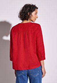 Camicia rossa leggera a maniche lunghe con colletto rotondo e dettaglio arricciato sul retro, abbinata a jeans in denim blu.
