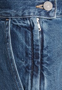Gros plan d’un jean en denim bleu montrant une braguette partiellement déboutonnée et un bouton en métal gravé "Ralph Lauren."
