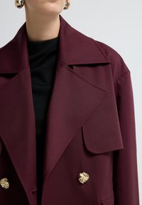 Bordeaux dobbeltbrystet jakke med store slag, prydet med guldfarvede løvehoveder knapper og en glat tekstur, båret over en sort bluse.