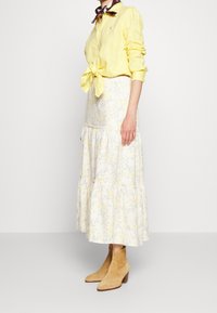 Keltainen pellavapaita, jossa on solmu vyötäröllä, vaaleista väreistä koostuva kukkakuvioinen kerroksellinen maksimekko ja vaaleat nahkasaappaat avokorkolla.