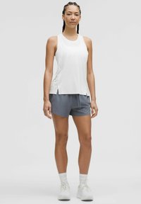Witte sportieve tanktop met zijsplitten, gecombineerd met grijze shorts. Model draagt witte sokken en bijpassende schoenen. Soepele stofstructuur.