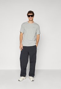 Mann trägt ein graues kurzärmliges Hemd, schwarze Cargo-Hosen, weiße Sneakers und große schwarze Sonnenbrillen, steht mit einer Hand in der Tasche.