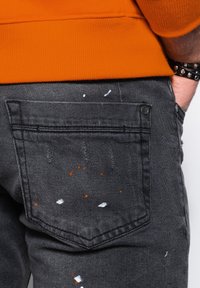 Harmaat farkut, joissa oranssin ja valkoisen maalitahroja. Takataskussa ompelu ja hopeinen napin yksityiskohta.