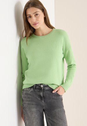 Maglione verde chiaro lavorato a maglia con scollo rotondo e maniche lunghe, abbinato a jeans grigio scuro. La texture appare morbida e casual.