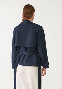 Námořnická modrá bunda s dlouhými rukávy, páskem v oblasti pasu a vrstvou na zadním panelu. Hladká tkanina s klasickým límcem a přizpůsobeným střihem.