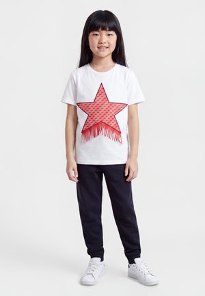 Jeune fille aux longs cheveux foncés portant un t-shirt blanc avec une grande étoile rouge et un pantalon noir, debout et souriante sur un fond clair.