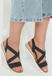 Mustat nahkasaappaat, joissa on ristikkäiset hihnat, matala korokepohja, jossa on luonnonkuitu-aksentti, ja valkoinen kumipohja.