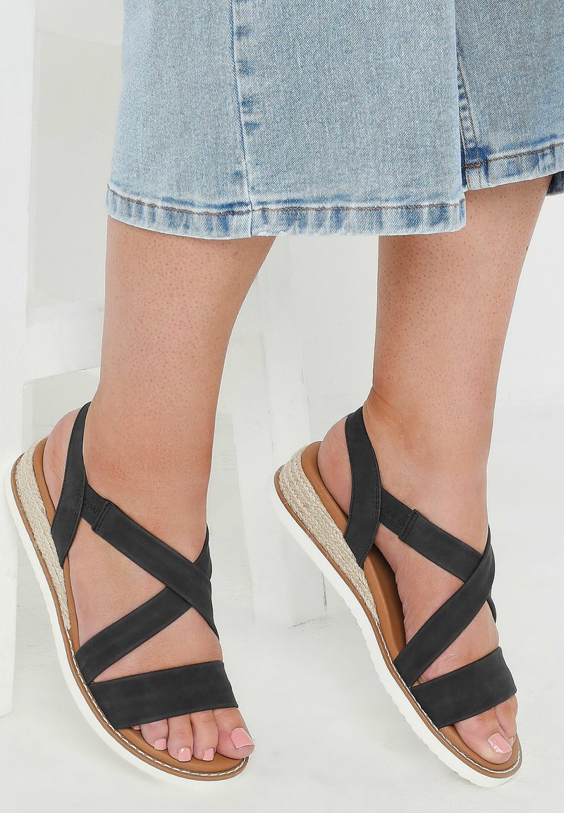 Mustat nahkasaappaat, joissa on ristikkäiset hihnat, matala korokepohja, jossa on luonnonkuitu-aksentti, ja valkoinen kumipohja.