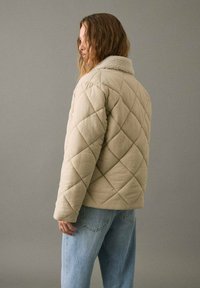 Mujer con cabello ondulado que lleva una chaqueta acolchada beige con cuello de sherpa, de pie de perfil sobre un fondo gris liso.