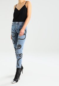 Vrouw in lichtblauwe jeans met zwarte abstracte patronen, een zwart mouwloos topje en zwarte hoge hakken met zilveren neuzen.