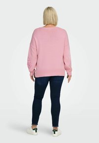 Maglione rosa oversize con orlo e polsini a coste, caratterizzato da un disegno floreale in rilievo sulla manica sinistra. Indossato con jeans scuri e sneakers bianche.