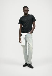 T-shirt preta com um pequeno logótipo branco, combinada com jeans de lavagem clara e sapatos pretos. Um hoodie branco está drapeado na cintura.