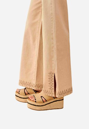 Pantalones beige de pierna ancha con bajos bordados y pequeñas aberturas laterales, usados con sandalias de plataforma tejidas con detalles en negro.