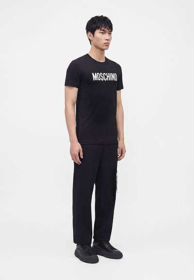 Άνδρας μοντέλο που φοράει μαύρο t-shirt Moschino, μαύρο παντελόνι και μαύρα αθλητικά παπούτσια, στέκεται μπροστά σε απλό λευκό φόντο.