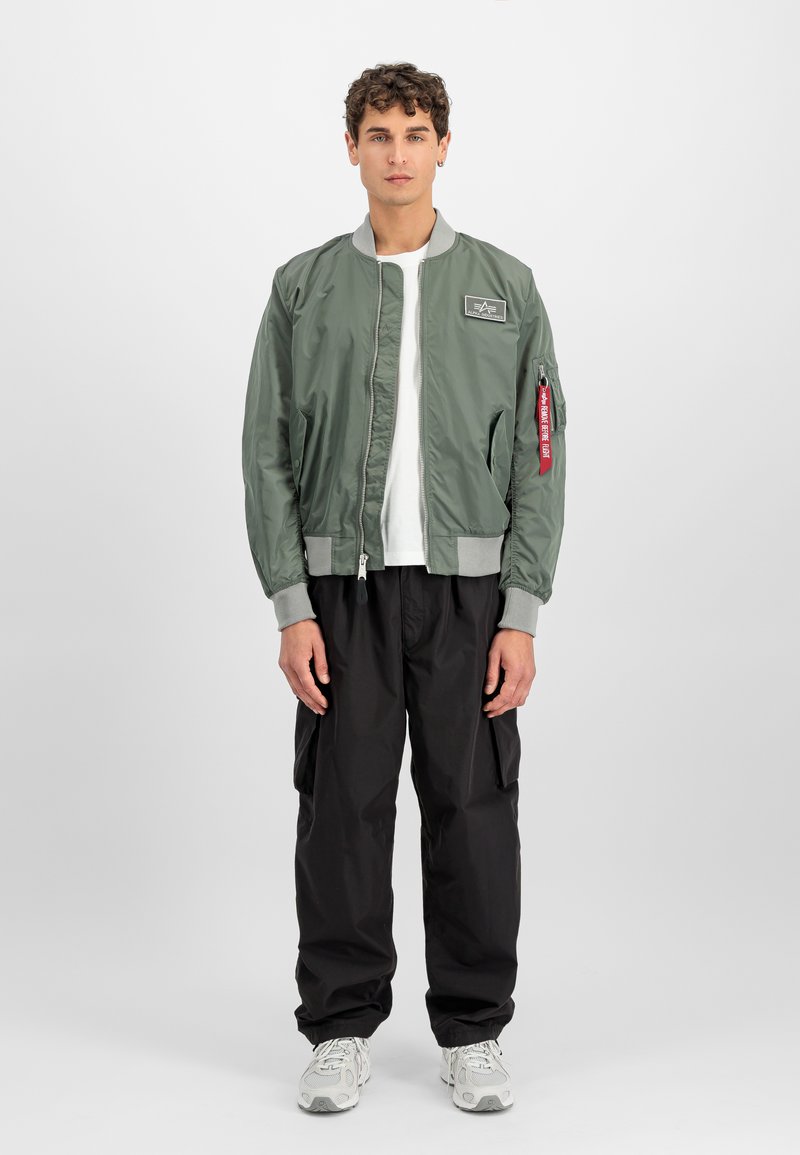 Sage olive bomber over a white T-shirt with black cargo pants showing large thigh pockets and white sneakers