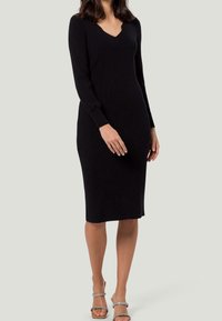 Robe en tricot noir à manches longues, décolleté en V, silhouette ajustée et longueur genou. Portée avec des sandales à lanières et des accessoires minimalistes.