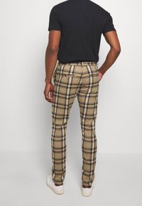 Beige plaid trousers with a slim fit, featuring black and white check patterns, two back pockets, and a smooth fabric texture. White sneakers visible.