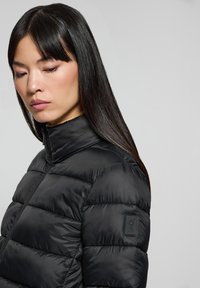 Giacca a vento nera con collo alto, caratterizzata da sezioni trapuntate e una piccola etichetta con logo sulla manica. Materiale liscio e lucido.