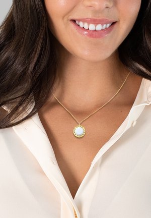 Collana d'oro con un pendente a scallop che presenta una pietra semi-traslucida di colore blu chiaro. Chi indossa la collana è vestita con una blusa chiara con bottoni.