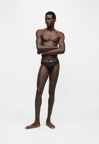 Homme torse nu, bras croisés, debout en slip noir avec motif grec doré à la ceinture, tourné légèrement vers la gauche sur un fond uni.