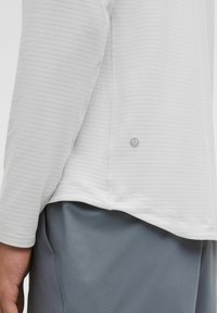 Koszulka sportowa z długim rękawem w jasnoszarym kolorze, z delikatnymi poziomymi paskami, z zaokrąglonym dołem i metką z logo na dolnej części pleców.