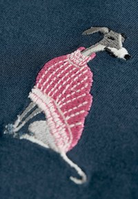 Broderad vinthund som bär en rosa tröja, sitter på en mörkblå tygbakgrund och visar detaljerad sömnad och design.