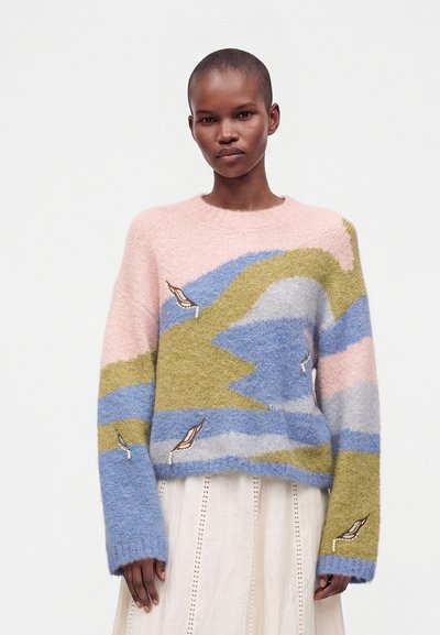 Personne portant un pull en peluche pastel rose, bleu, vert et gris avec une broderie d'oiseau, associé à une jupe blanche sur un fond uni.