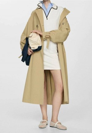 Trench-coat beige ceinturé à la taille, porté sur une robe blanche et une chemise bleue à col. Chaussures beiges avec une bride transparente. Porte un sac.