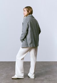 Mujer con blazer de lana gris, pantalones de pierna ancha blancos y zapatos beige, de pie de lado con las manos en los bolsillos contra un fondo sencillo.