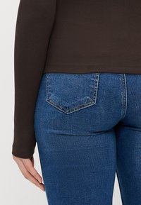 Camisa de manga longa castanha canelada, metida dentro de jeans azuis de cintura alta. Os jeans apresentam um design clássico de cinco bolsos, com um leve desbotado e um tecido texturizado.