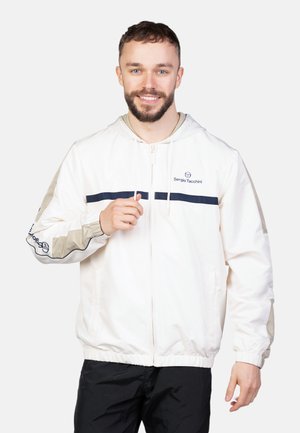 Leichte Cremefarbe Jacke mit einer Kapuze, die marineblaue Streifen auf der Brust und den Ärmeln aufweist, elastische Bündchen und einen Reißverschluss. Logo-Detail sichtbar.