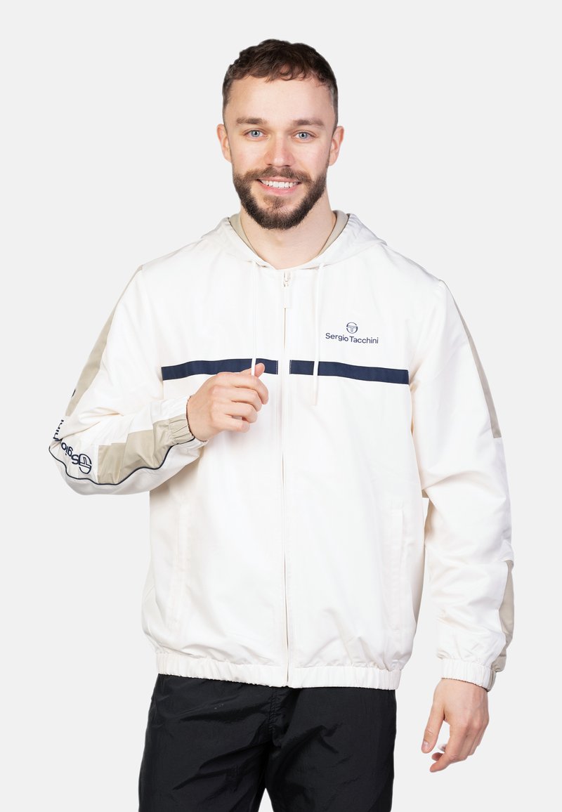 Chaqueta ligera de crema con capucha, con rayas marinas en el pecho y las mangas, puños elásticos y cierre de cremallera. Detalle del logotipo visible.