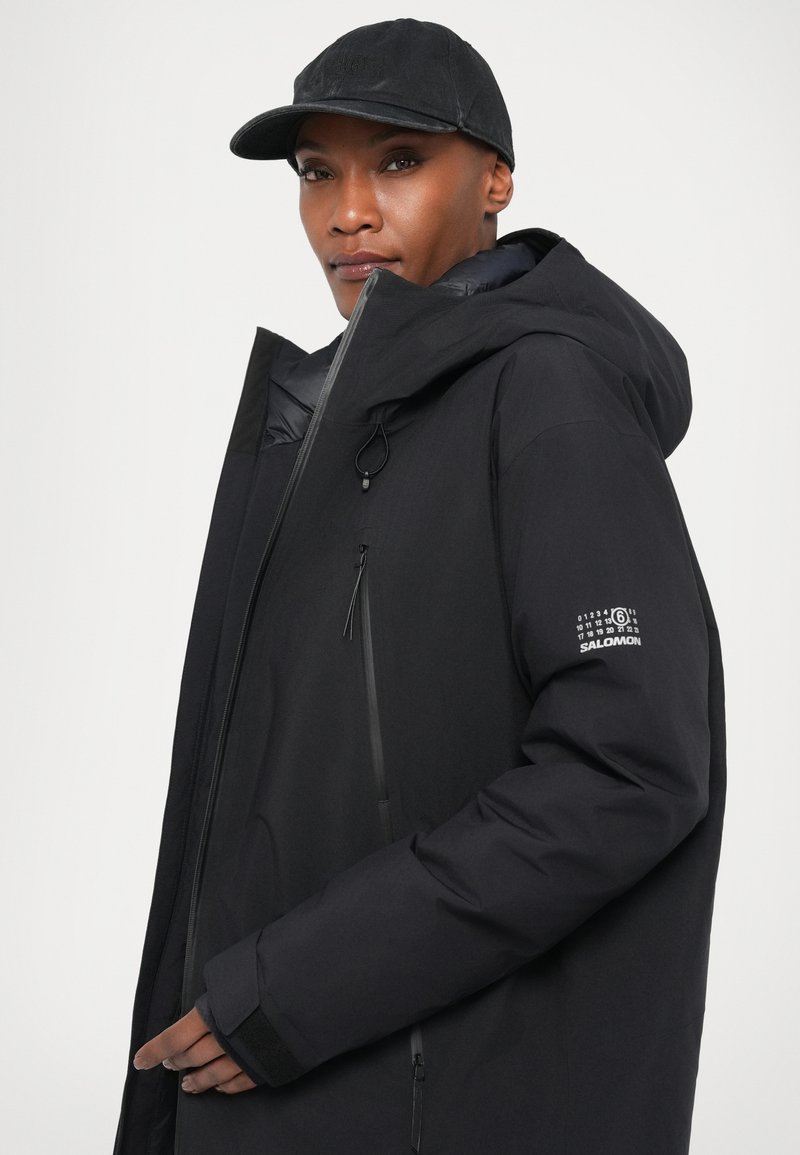 Veste isolante noire avec un col haut et une capuche ajustable, dotée de poches zippées et d'un logo sur la manche. Fini lisse et mat.