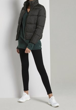 Giacca a vento nera con colletto alto, abbinata a un maglione di lana teal, jeans skinny neri e scarpe da ginnastica bianche.