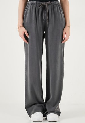 Naine, kes kannab lahtisi halli nööriga pükse ja valgeid tossusid, seisab käed külgede lähedal lõdvestunult tavalise tausta ees.